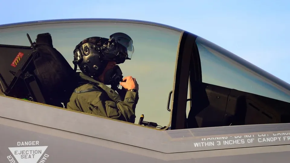 Un pilot cu cască de zbor și mască de oxigen stă în cockpitul unui avion sub un cer senin.