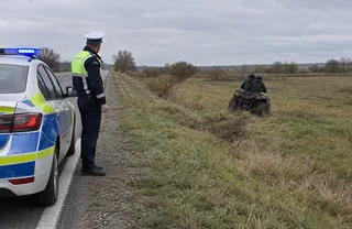 Un polițist stă pe marginea drumului lângă o mașină de serviciu, privind spre un ATV pe care se află două persoane într-un câmp.