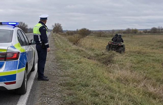 Un polițist stă pe marginea drumului lângă o mașină de serviciu, privind spre un ATV pe care se află două persoane într-un câmp.