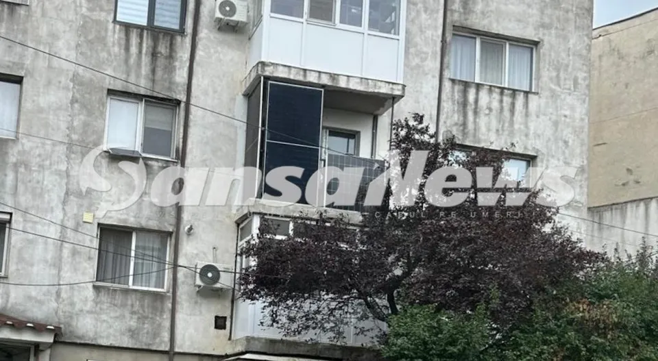 Panouri fotovoltaice negre sunt montate pe balconul unui apartament de la un etaj al unei clădiri gri cu ferestre și un arbore în prim-plan.