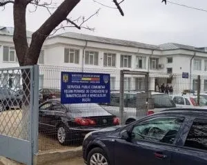 O clădire albă cu ferestre este vizibilă în spatele unui gard metalic și a mai multor mașini parcate pe stradă.