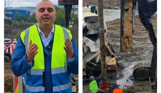 Un bărbat cu vestă reflectorizantă galbenă și albastră stă în fața unui șantier, lângă utilaje de construcții și pământ excavat.