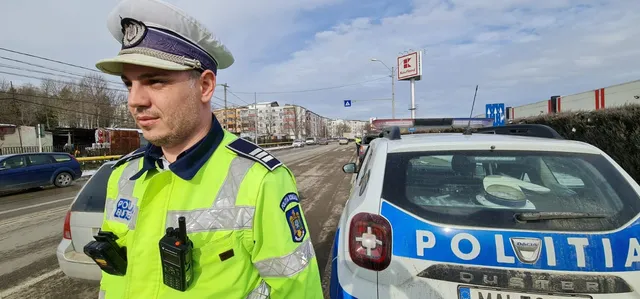 Un polițist în uniformă reflectorizantă stă lângă o mașină de serviciu albastră și albă pe un drum asfaltat sub un cer înnorat.