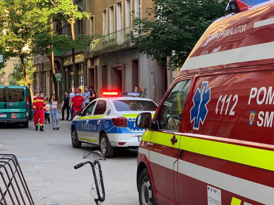 O ambulanță roșie și o mașină de poliție alb-albastră cu girofaruri aprinse pe o stradă, cu oameni în fundal.