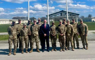 Un grup de soldați în uniforme de camuflaj stau alături de un bărbat în costum întunecat și cravată roșie pe un teren deschis sub un cer înnorat.