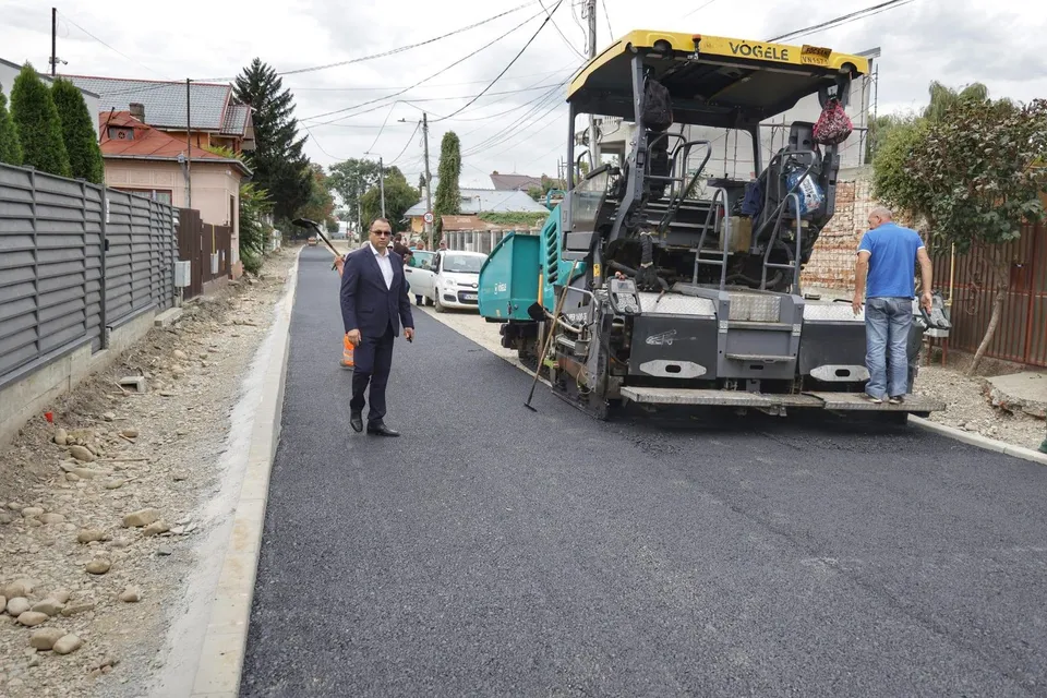 Un bărbat în costum merge pe asfalt proaspăt, lângă o autofinalizatoare și alți doi bărbați, pe o stradă cu case și garduri.