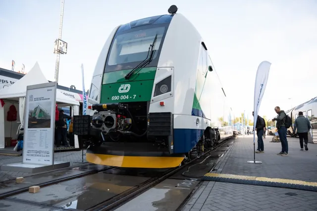 Un tren alb, verde și albastru stă pe șine lângă un panou informativ și câțiva oameni pe o platformă pavată.