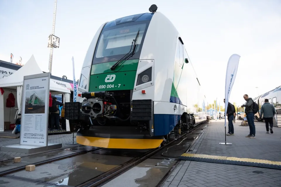 Un tren alb, verde și albastru stă pe șine lângă un panou informativ și câțiva oameni pe o platformă pavată.
