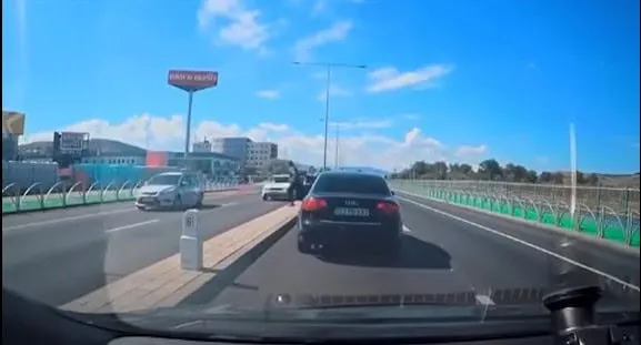 Privire de pe parbrizul unui autovehicul către un drum cu mai multe benzi, pe care circulă mașini sub un cer senin.