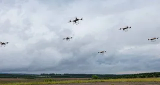 Șase drone mici zboară pe un cer acoperit de nori deasupra unui câmp deschis cu vegetație la orizont.