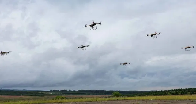 Cinci drone mici zboară pe un cer acoperit de nori deasupra unui peisaj de câmpie cu vegetație verde și copaci în depărtare.