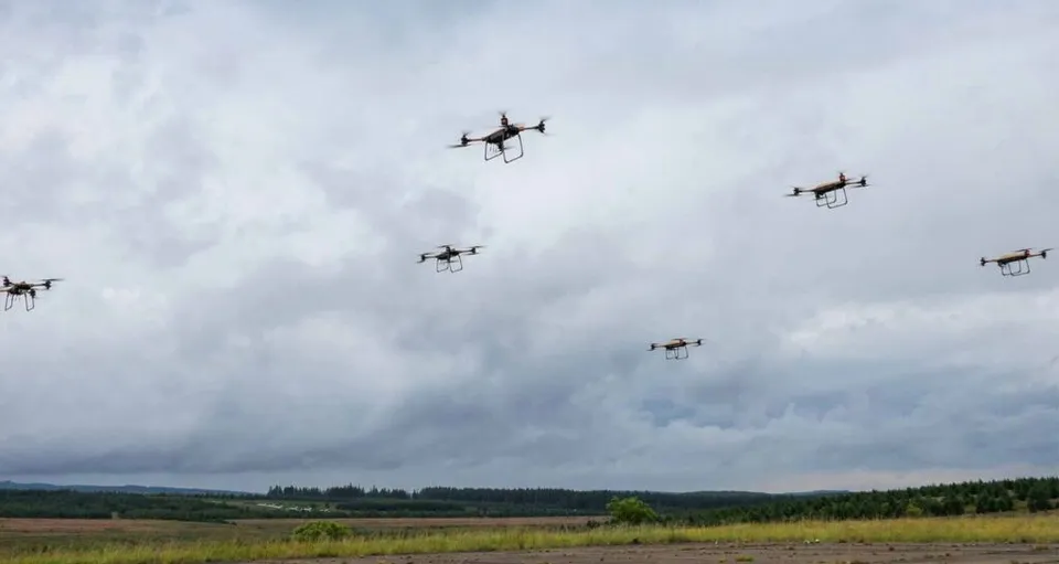 Cinci drone mici zboară pe un cer acoperit de nori deasupra unui peisaj de câmpie cu vegetație verde și copaci în depărtare.