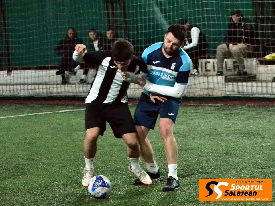 Doi bărbați în echipamente sportive se luptă pentru un minge de fotbal pe un teren artificial, cu un șir de spectatori în fundal.