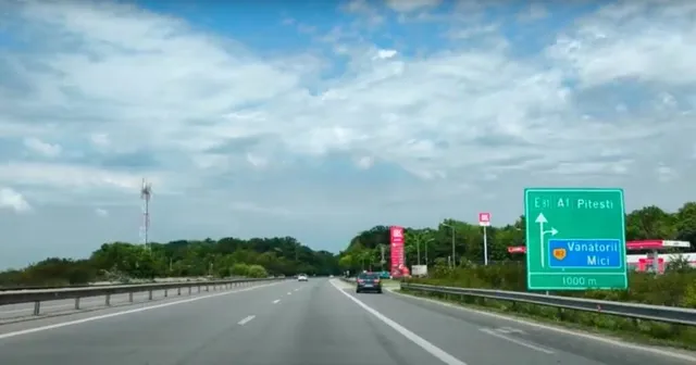 O autostradă cu două benzi de rulare se întinde spre orizont sub un cer cu nori, având un panou indicativ verde în partea dreaptă.