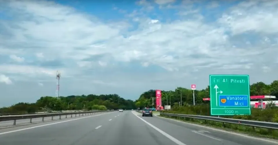 O autostradă cu două benzi de rulare se întinde spre orizont sub un cer cu nori, având un panou indicativ verde în partea dreaptă.