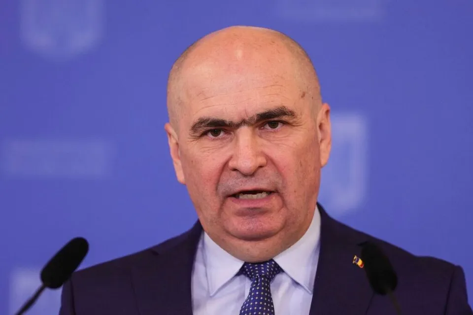 Un bărbat cu părul rare și costum închis la culoare vorbește în fața a două microfoane pe un fundal albastru.