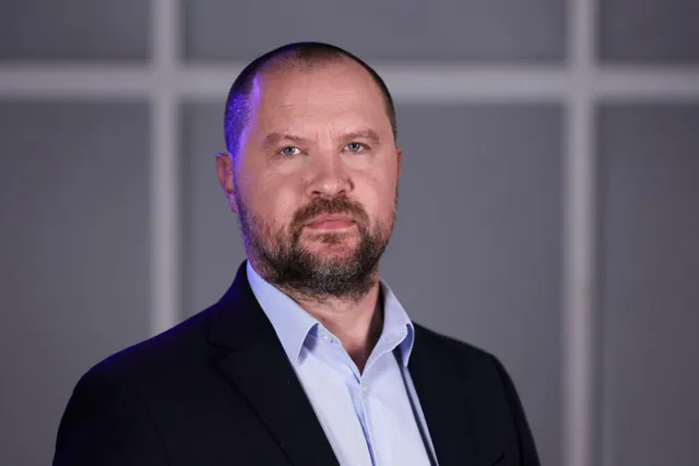 Un bărbat cu barbă și păr scurt, purtând o cămașă deschisă la guler și un sacou închis la culoare, privește spre cameră pe un fundal gri.