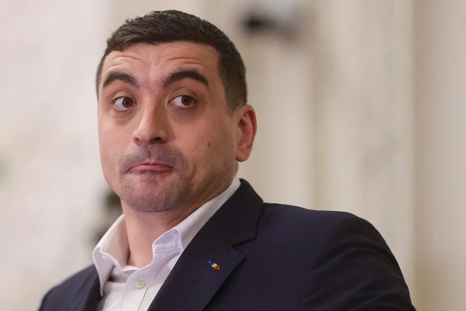 Un bărbat cu părul scurt și scurtă barbă poartă o cămașă albă și un sacou închis la culoare cu un pin pe rever.