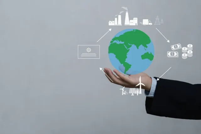 O mână întinsă susține o ilustrație digitală a globului pământesc înconjurat de pictograme albe reprezentând industrie, bani, energie și documente pe un fundal gri.