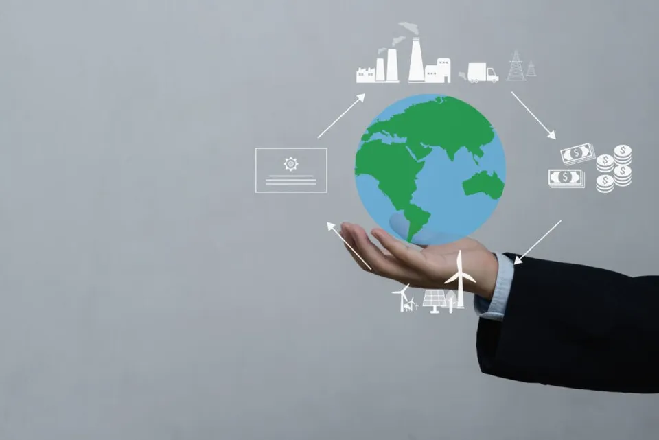 O mână întinsă susține o ilustrație digitală a globului pământesc înconjurat de pictograme albe reprezentând industrie, bani, energie și documente pe un fundal gri.