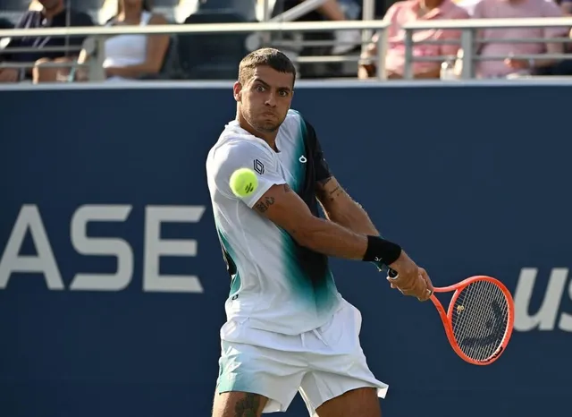 Un bărbat cu tricou alb și pantaloni scurți de sport lovește o minge de tenis cu o rachetă portocalie pe un teren de tenis.