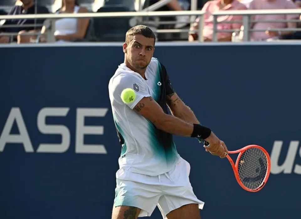 Un bărbat cu tricou alb și pantaloni scurți de sport lovește o minge de tenis cu o rachetă portocalie pe un teren de tenis.