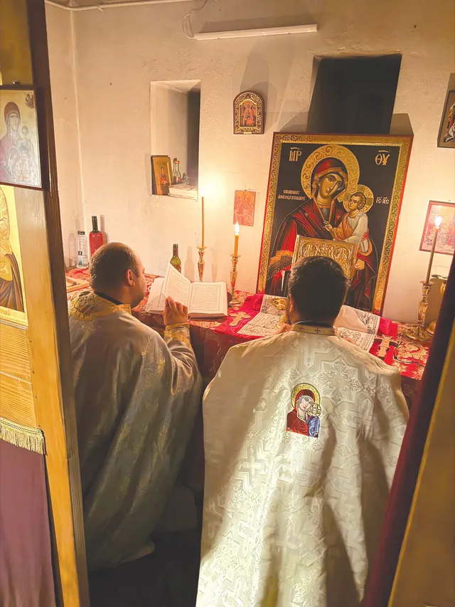 Doi bărbați în haine liturgice stau în fața unui altar pe care se află o icoană mare și o carte deschisă.