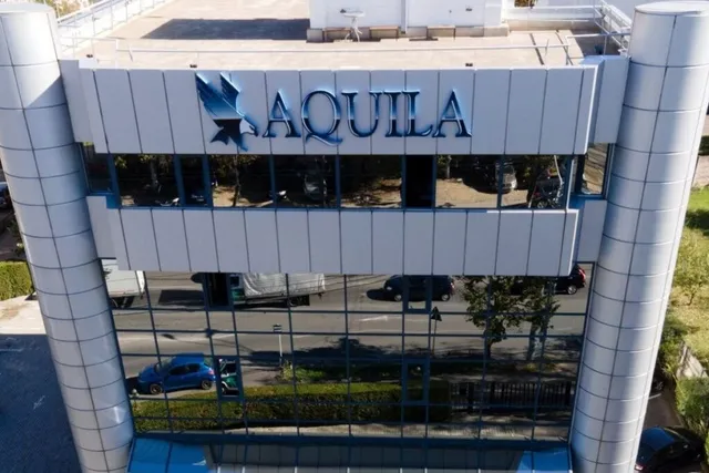 O clădire cu fațadă de sticlă și panouri gri prezintă un logo albastru cu textul AQUILA deasupra ferestrelor întunecate.