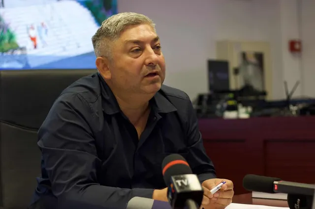 Un bărbat cu părul scurt și cămașă închisă stă la o masă în fața mai multor microfoane.