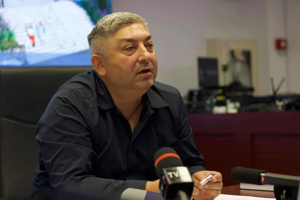 Un bărbat cu părul scurt și cămașă închisă stă la o masă în fața mai multor microfoane.
