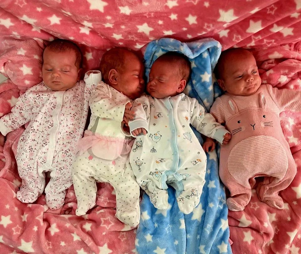 Patru nou-născuți îmbrăcați în pijama de culcare stau întinși pe o pătură roz cu motive stelare.