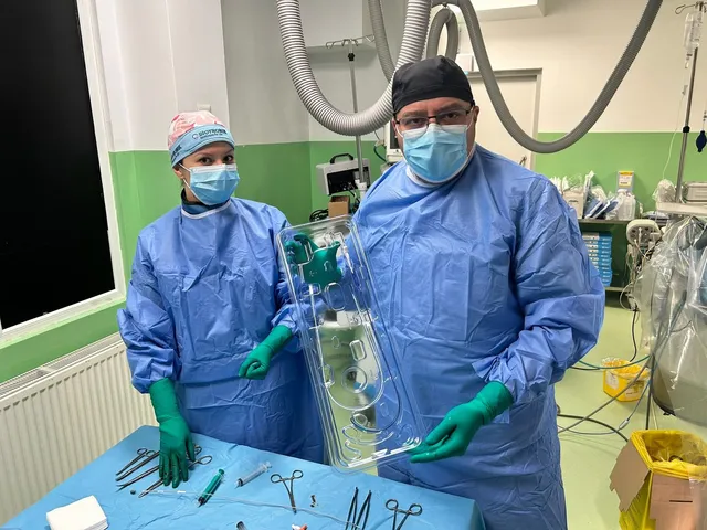 Un bărbat și o femeie în halate medicale albastre și măști de protecție țin un recipient transparent de plastic deasupra unei mese cu instrumente chirurgicale metalice.