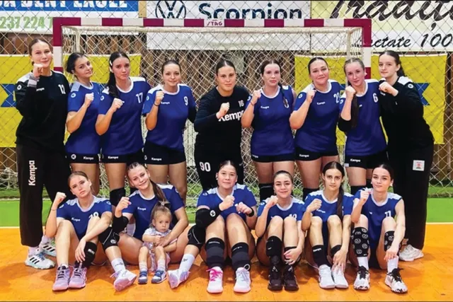 O echipă de handbal feminin posedă jucătoarele în poziții diverse, unele stând în picioare și altele așezate pe un teren de culoare portocalie.