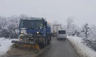 Un camion albastru cu echipament de deszăpezire circulă pe un drum îngust acoperit de zăpadă, urmat de un van alb și un camion alb.
