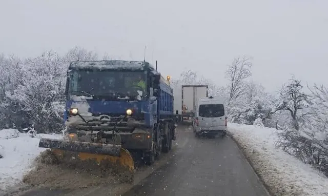 Un camion albastru cu echipament de deszăpezire circulă pe un drum îngust acoperit de zăpadă, urmat de un van alb și un camion alb.
