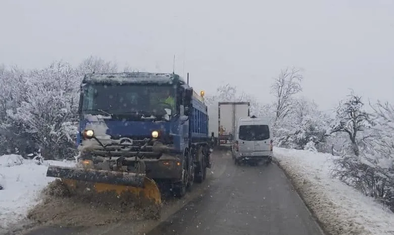 Un camion albastru cu echipament de deszăpezire circulă pe un drum îngust acoperit de zăpadă, urmat de un van alb și un camion alb.