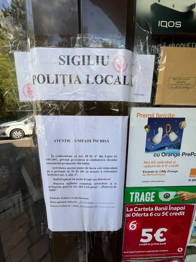 Un anunț alb pe o suprafață transparentă afișează text negru și un sigiliu, fiind lipit pe o vitrină de magazin lângă diverse reclame colorate.