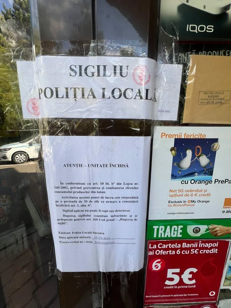 Un anunț alb pe o suprafață transparentă afișează text negru și un sigiliu, fiind lipit pe o vitrină de magazin lângă diverse reclame colorate.