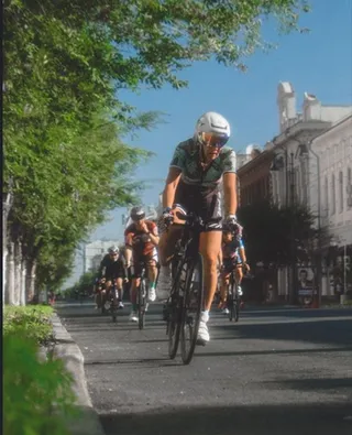 Mai mulți cicliști în echipament sportiv pedalează pe un drum asfaltat lângă un trotuar cu copaci și clădiri în fundal.