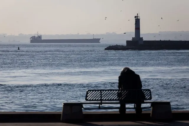 O persoană stă pe o bancă de lângă apă, cu un vapor și un far în fundal.
