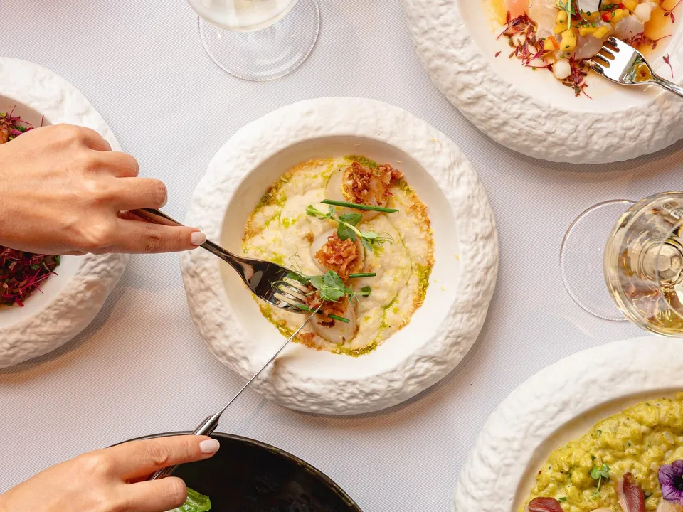 Mâini care folosesc furculițe pentru a mânca preparate culinare din farfurii albe, texturate, așezate pe o masă cu față albă.