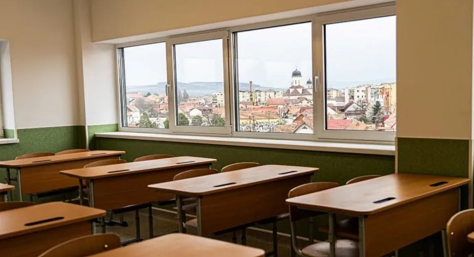 Mai multe bănci de lemn cu picioare metalice sunt așezate într-o sală de clasă lângă o fereastră lată cu vedere spre un oraș.
