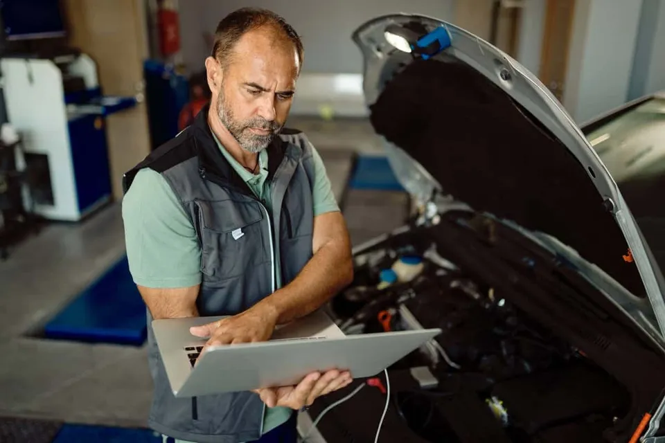 Un bărbat cu vestă gri și tricou deschis consultă un laptop în timp ce se află lângă un motor de mașină cu capota deschisă.