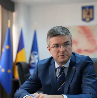 Un bărbat cu ochelari și costum albastru stă așezat în fața unor steaguri într-o încăpere.