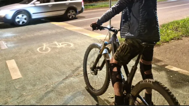 O persoană purtând pantaloni scurți și o jachetă neagră stă pe o bicicletă lângă o pistă marcată cu un simbol de bicicletă pe asfalt.