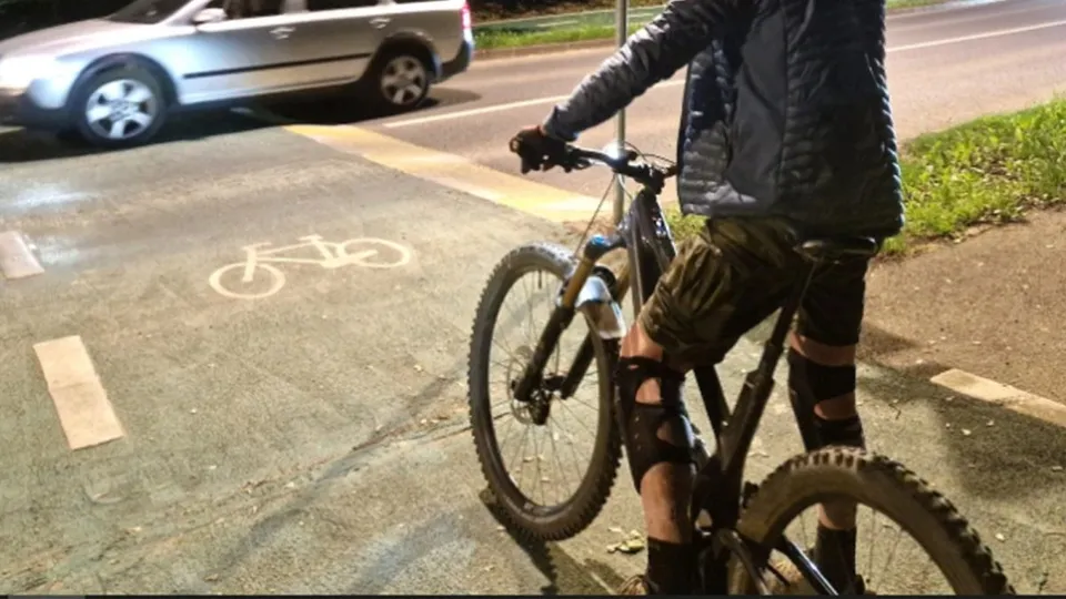 O persoană purtând pantaloni scurți și o jachetă neagră stă pe o bicicletă lângă o pistă marcată cu un simbol de bicicletă pe asfalt.