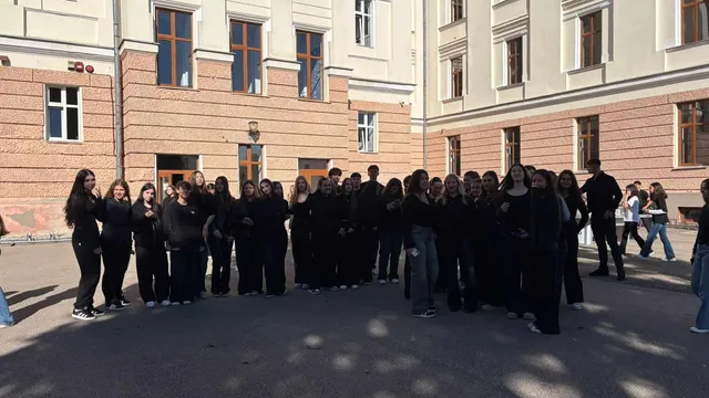 Un grup de persoane îmbrăcate în haine negre stă în fața unei clădiri cu ferestre mari și pereți din cărămidă și vopsea deschisă.