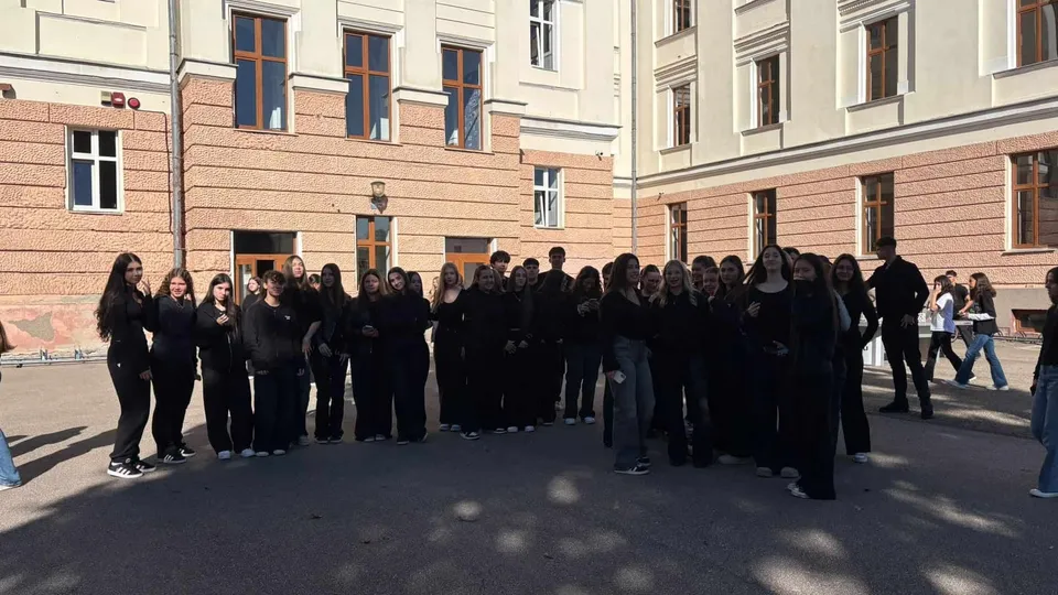 Un grup de persoane îmbrăcate în haine negre stă în fața unei clădiri cu ferestre mari și pereți din cărămidă și vopsea deschisă.