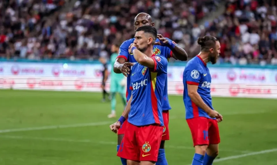 Trei jucători de fotbal în echipamente albastre și roșii stau pe un teren de fotbal verde în fața unei tribune cu spectatori.