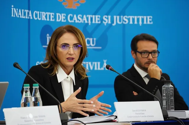 O femeie cu ochelari și o cămașă albă vorbește la un microfon, în timp ce un bărbat cu ochelari stă lângă ea în fundal.